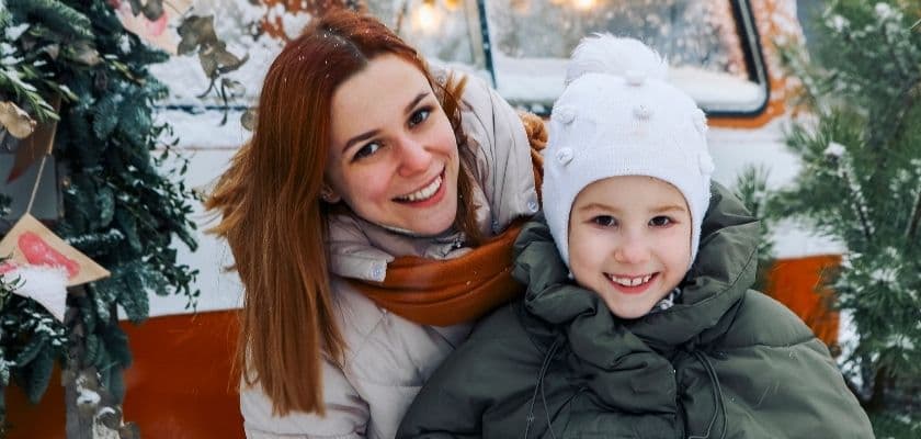 A smiling woman and child pose together outdoors in a snowy winter setting.