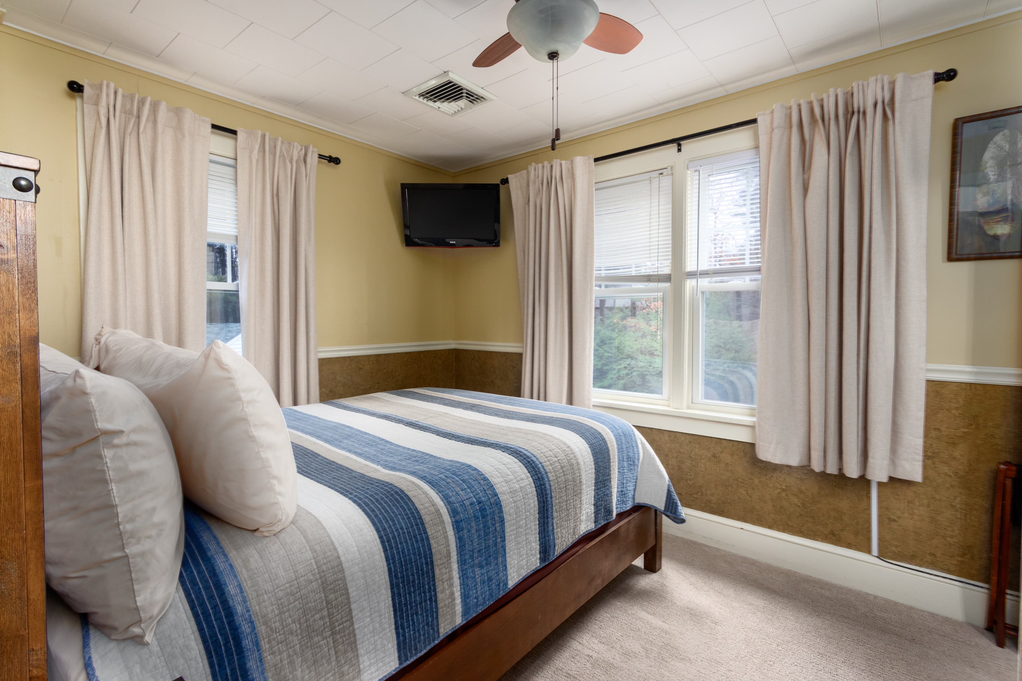 A bright bedroom with a dark wood bed, a blue and white striped quilt, pale yellow walls, and a wall-mounted TV between two windows.