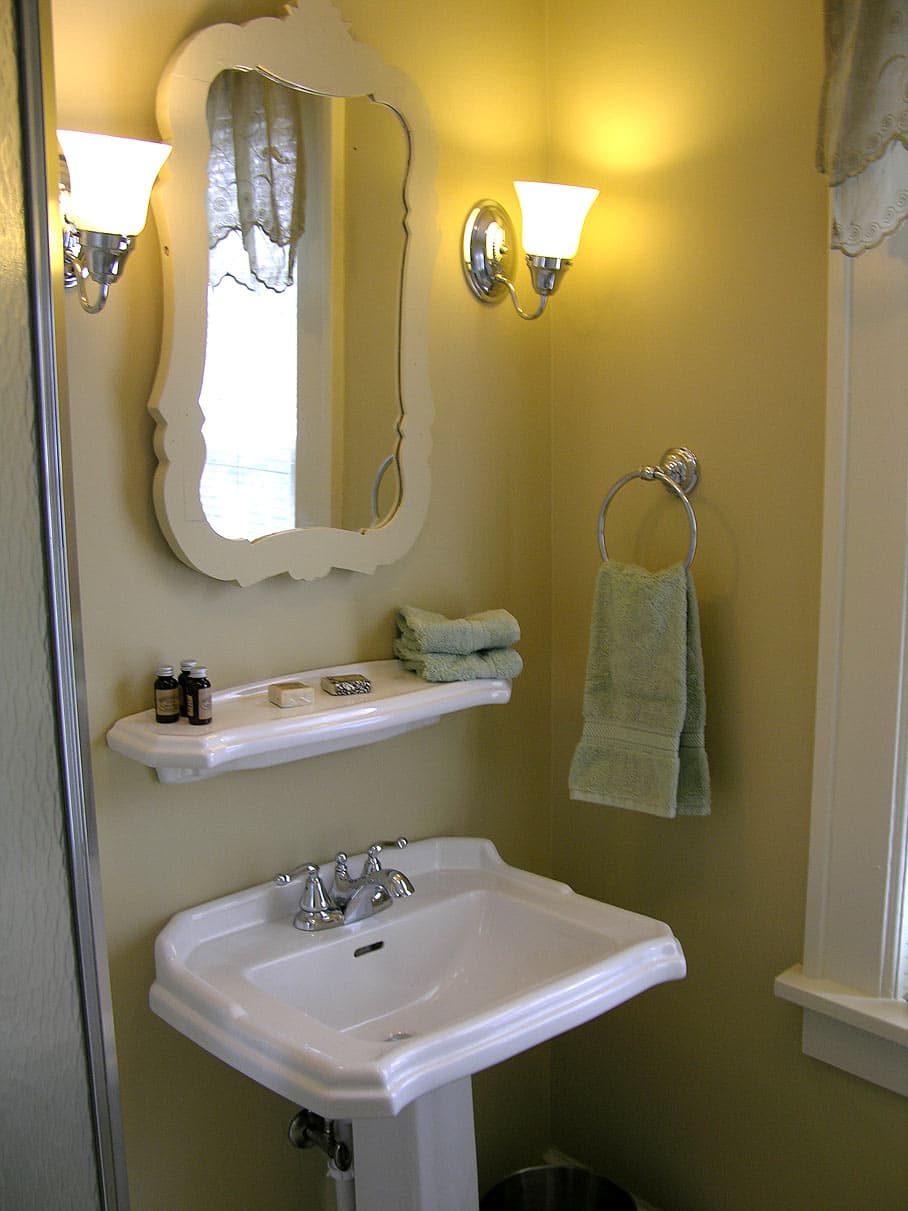 A charming bathroom vanity area with a white pedestal sink, a scalloped mirror, a small shelf, and sconce lighting on a pale yellow wall.