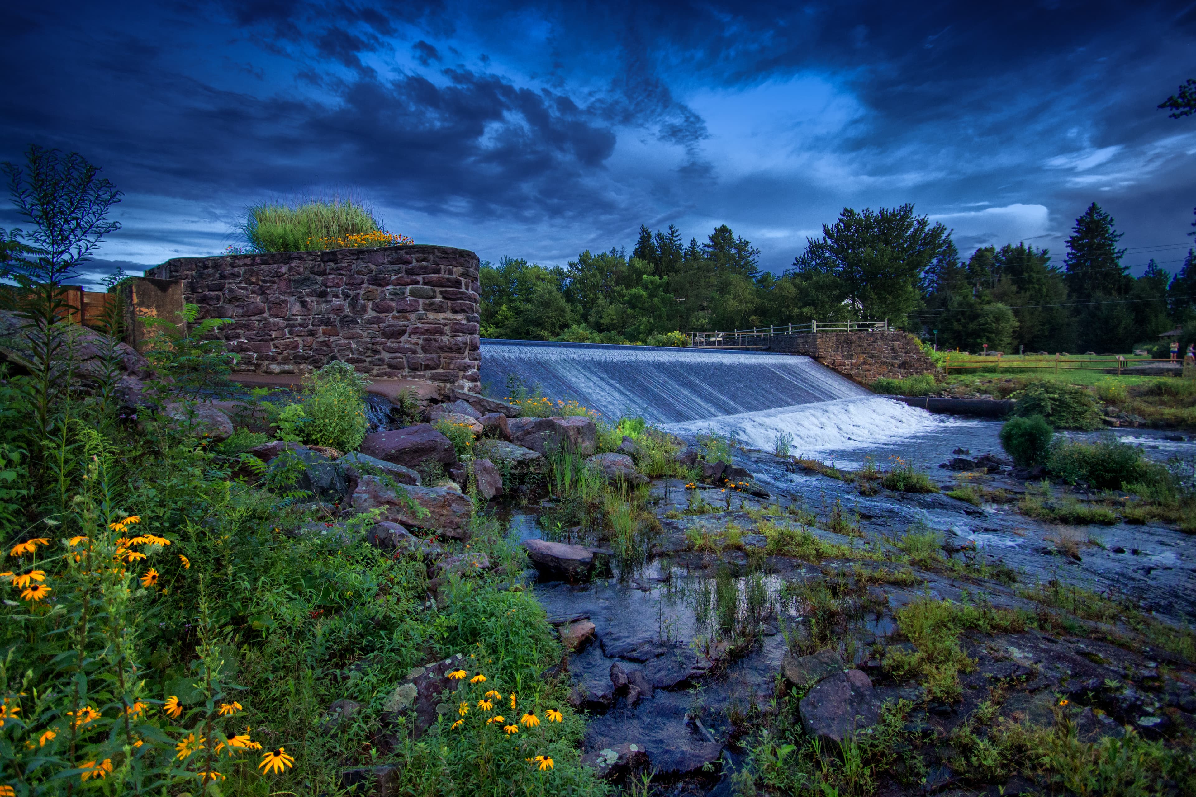 A stone dam with water cascading over it, surrounded by lush greenery and wildflowers under a moody sky.