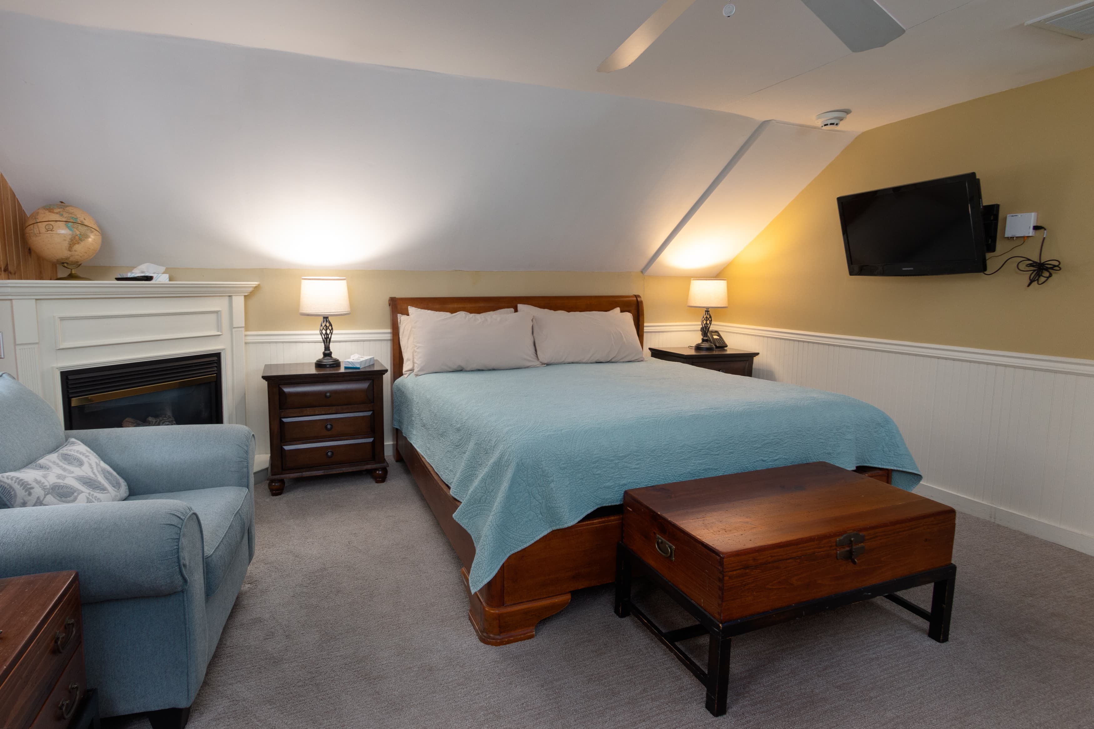 A ;third-floor bedroom with sloping ceilings, a king bed with a light blue quilt, a fireplace with a white mantle, and a wall-mounted TV.