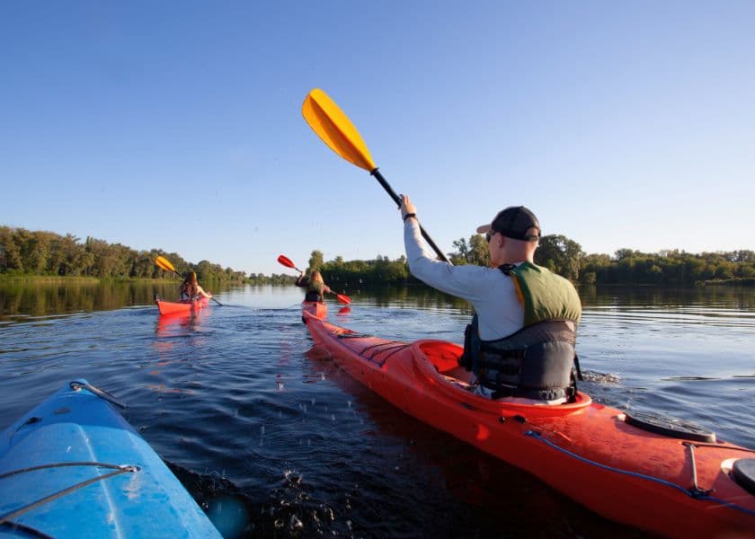 Three kayakers paddle on a calm river under a clear blue sky.