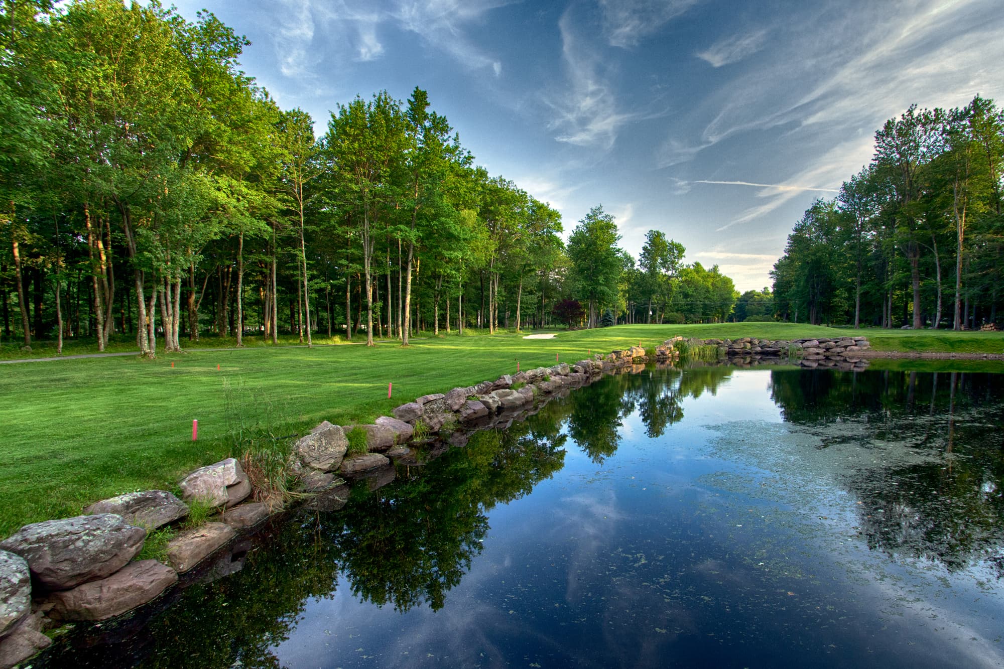 A serene golf course landscape with lush trees and a reflecting pond under a cloudy sky.