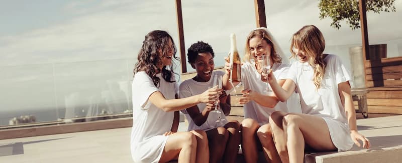 Four women in white t-shirts celebrate with drinks on a sunny patio.