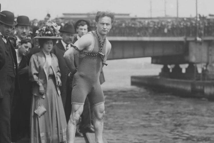A man in a swimsuit stands at the edge of a dock, bound by chains, while a crowd watches from the shore.