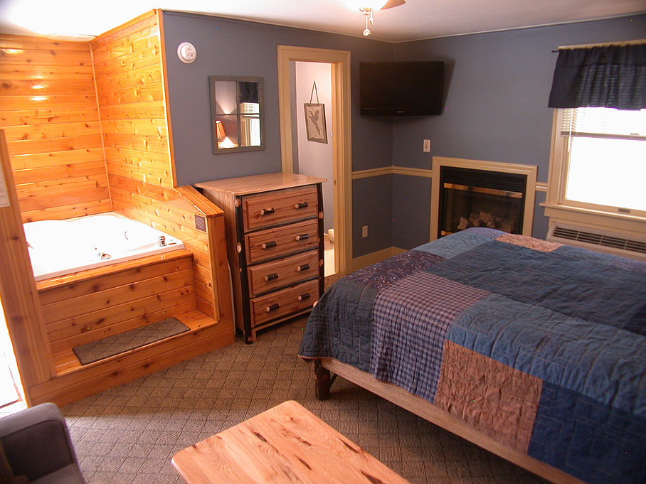 A rustic suite with a queen bed and a patchwork quilt, featuring a jetted tub in a wood-paneled corner, a fireplace, and a TV.