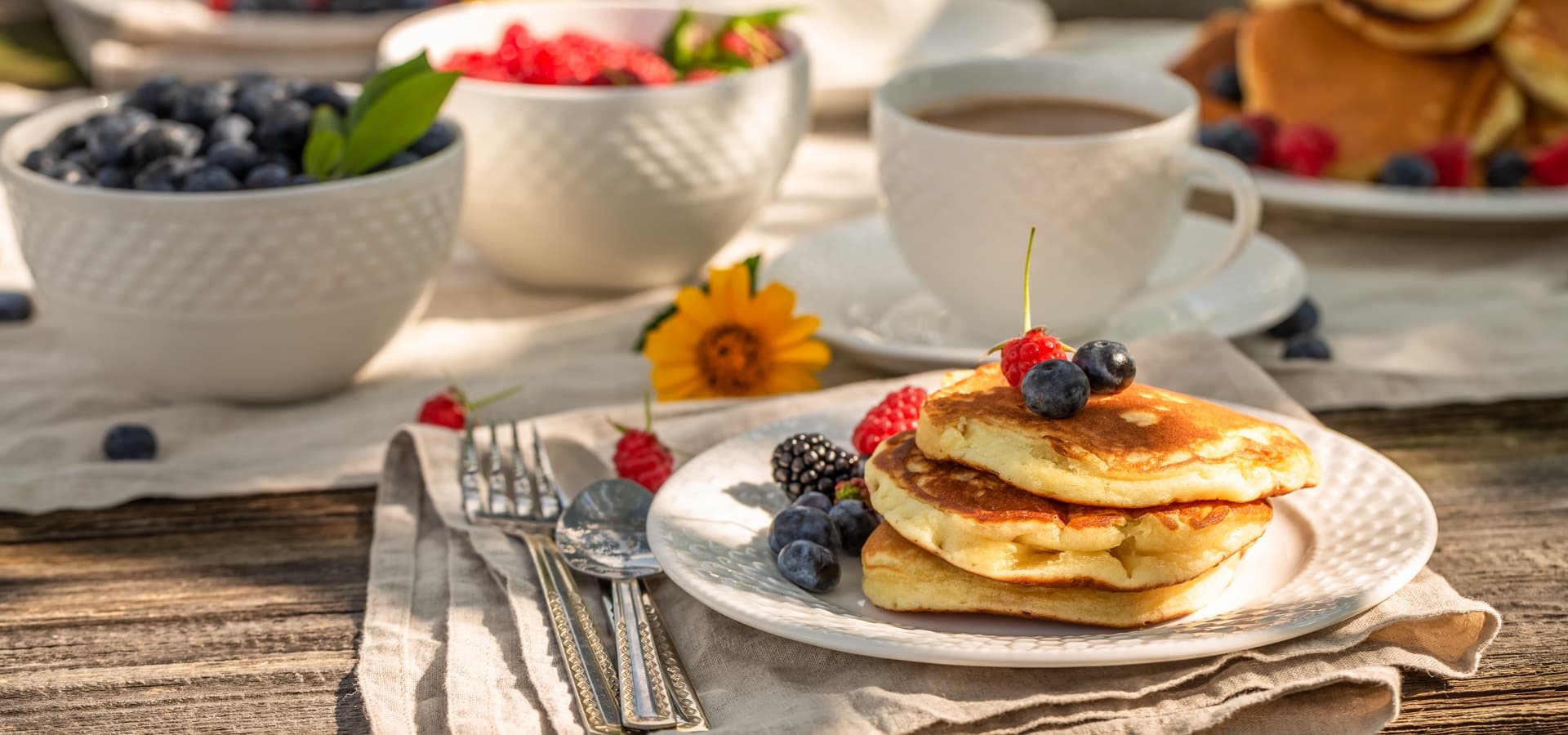 A plate of fluffy pancakes topped with berries sits on a wooden table beside a cup of coffee and bowls of fresh fruit.