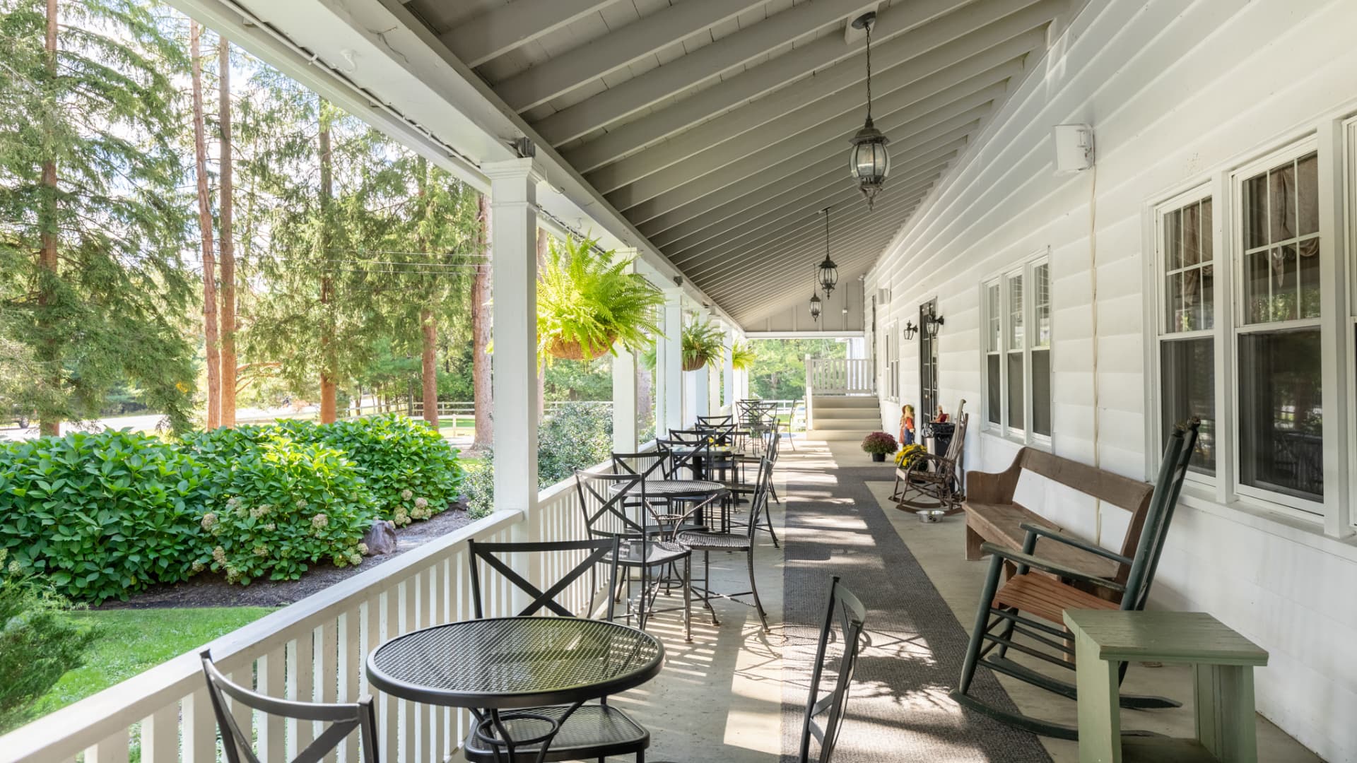 A cozy porch with black metal tables, rocking chairs, and hanging ferns, framed by lush greenery.