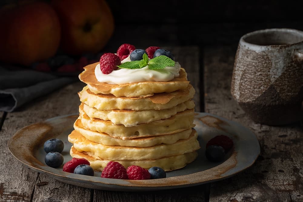 A stack of pancakes topped with cream, fresh raspberries, blueberries, and mint, served on a rustic plate.