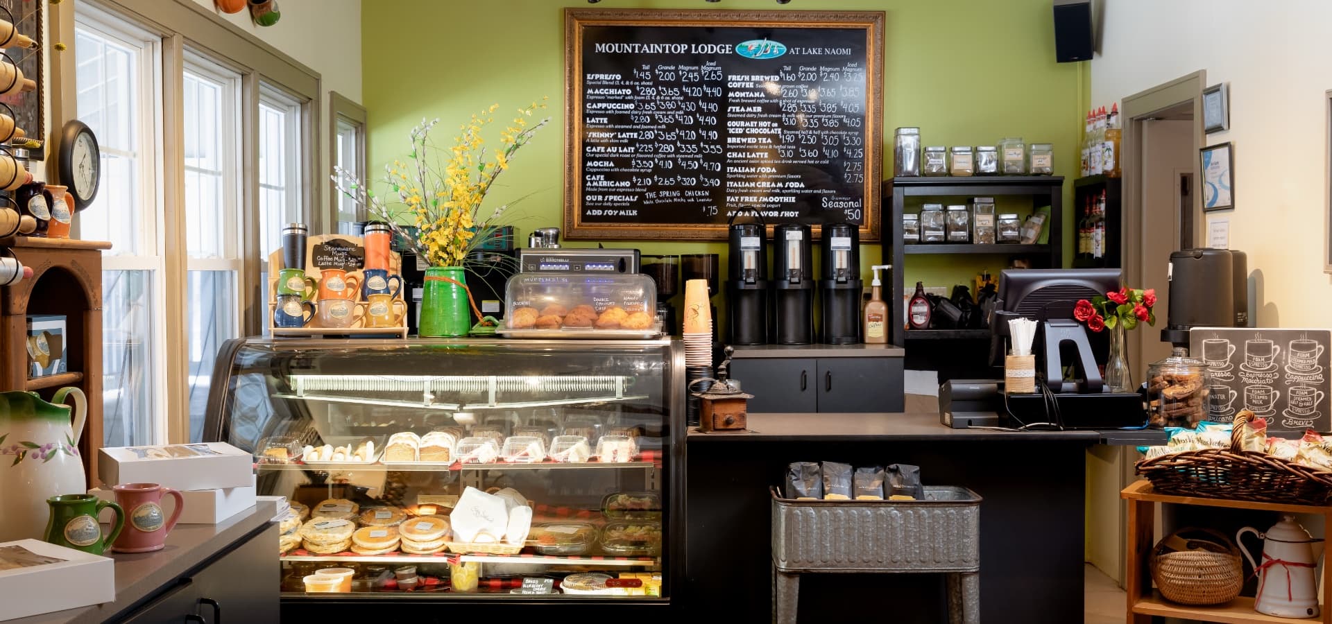 A cozy café interior featuring a display of pastries, a coffee menu, and decorative plants.