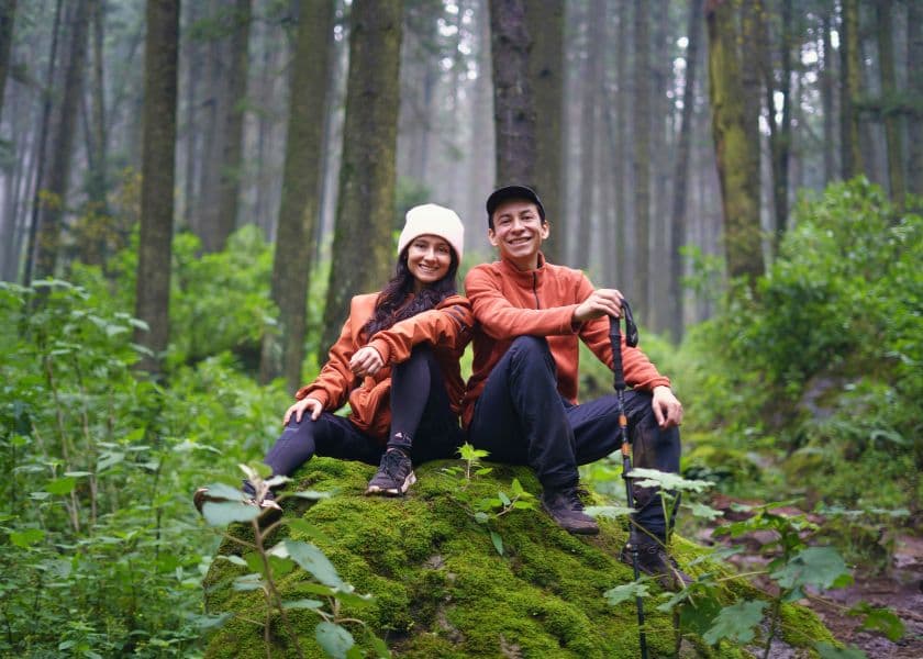 A couple relaxes on a moss-covered rock during a forest hike, surrounded by tall trees and lush greenery.