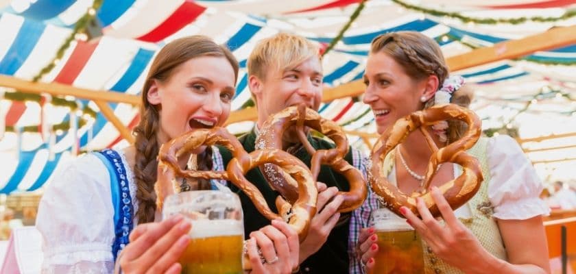 Three friends in traditional Bavarian outfits holding large pretzels and beers under festive striped bunting.
