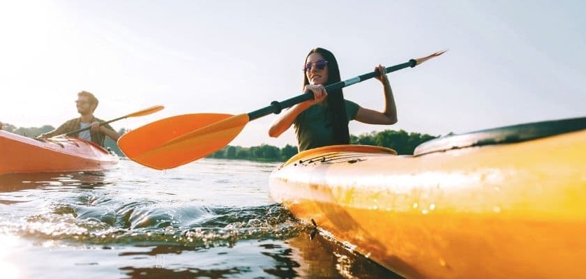 woman in sunglasses kayaking on a calm lake with sunlight reflecting off the water while a man paddles behind her in another kayak