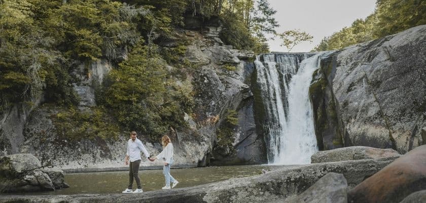 couple holding hands walking across rocks with a stony waterfall behind them