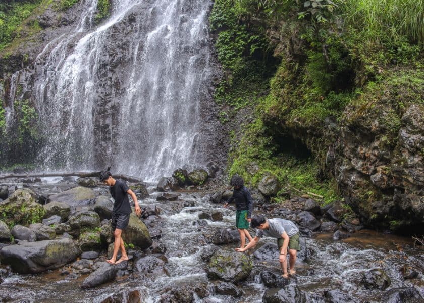 Three guys walking on rocks in a stream beneath a large waterfall surrounded by greenery